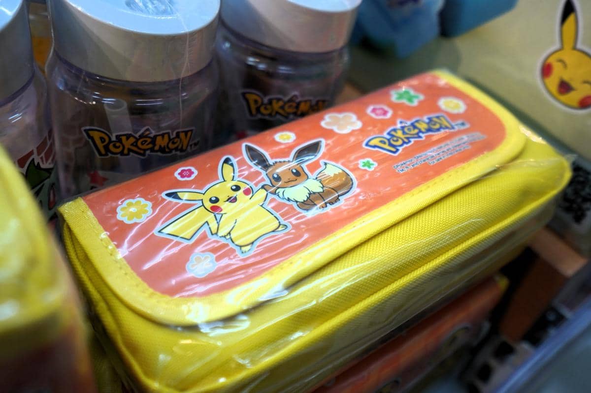 Tempat pensil berwarna kuning bergambar karakter Pokémon Pikachu dan Eevee, dipajang bersama botol minum bertema Pokémon di toko.