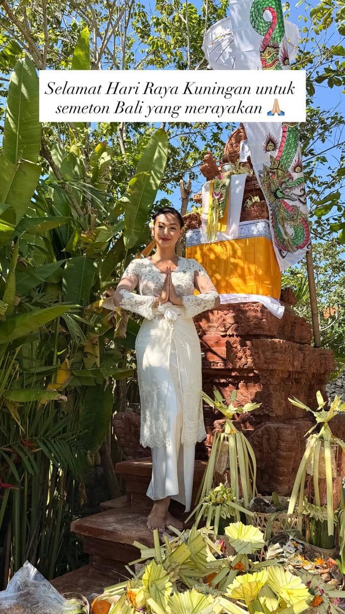 Indah  mengenakan kebaya putih berpose dengan tangan sembah di depan pelinggih berhias kain kuning saat perayaan Hari Raya Kuningan di Bali.