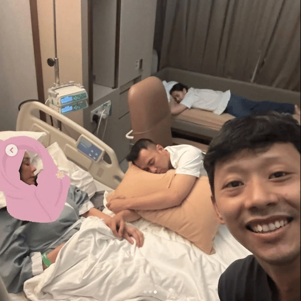 Seorang pria tersenyum di depan kamera di kamar rumah sakit, sementara dua orang lainnya tampak beristirahat di dekat tempat tidur pasien.
