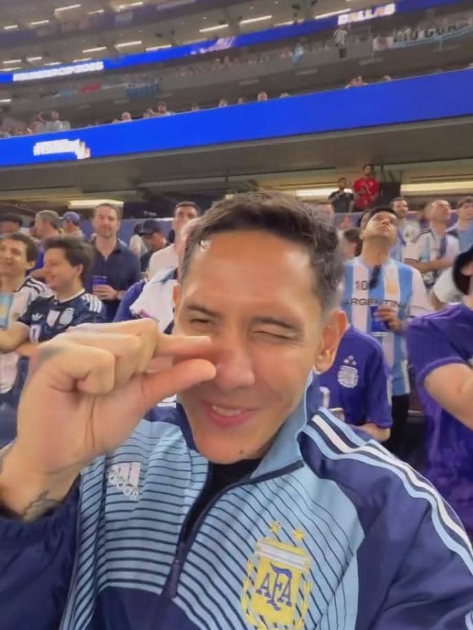 Seorang pria mengenakan jaket tim nasional Argentina tersenyum dan berpose di stadion yang dipenuhi penonton berpakaian serupa.