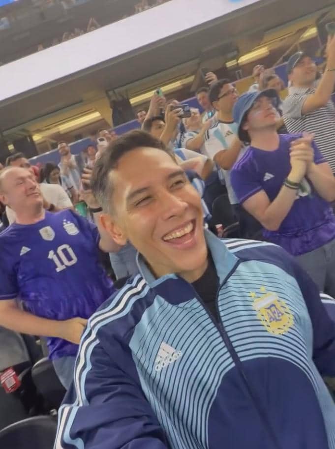 Seorang pria mengenakan jaket tim nasional Argentina tersenyum di stadion bersama para penonton yang mengenakan jersey Argentina.
