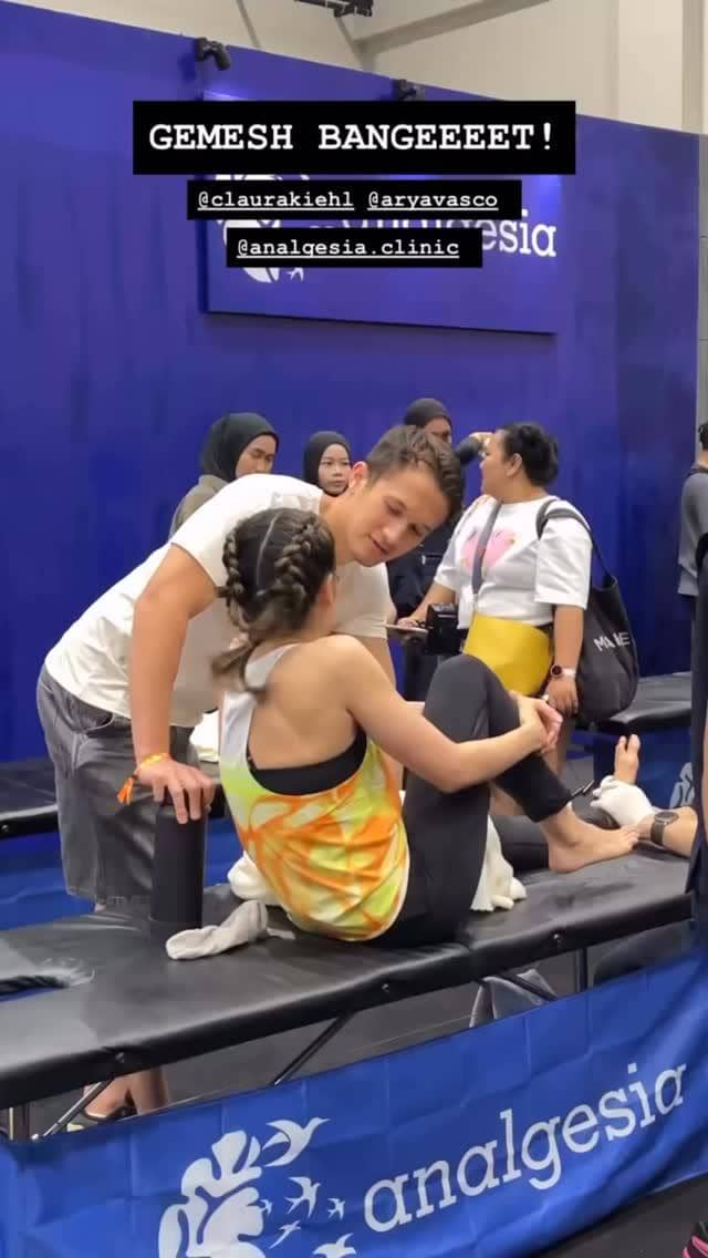 Seorang pria dan wanita berbincang akrab di area pijat olahraga dengan latar biru bertuliskan analgesia pada acara HYROX Jakarta 2026 Day 2.