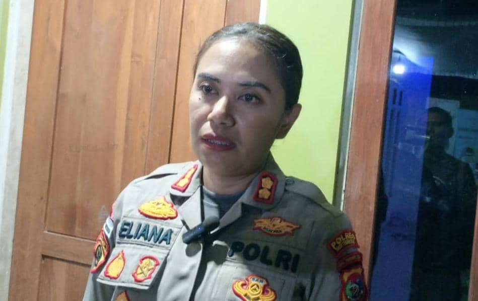 Seorang perwira polisi berpakaian dinas lengkap berdiri di depan pintu kayu dan kaca dengan ekspresi serius di dalam ruangan.