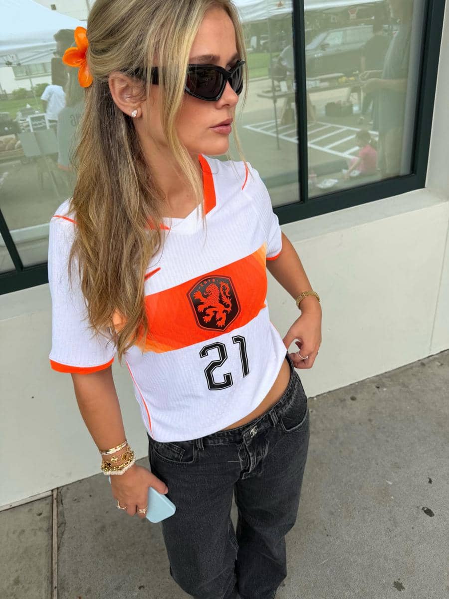Seorang perempuan mengenakan jersey tim nasional Belanda berwarna putih dan oranye dengan nomor 21, memakai kacamata hitam dan aksesori rambut oranye.