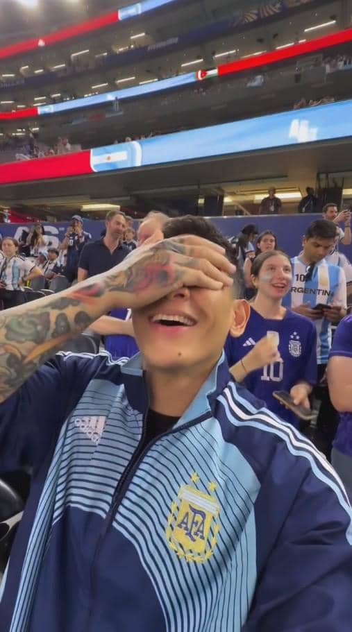Seorang pria mengenakan jaket tim nasional Argentina tersenyum sambil menutupi wajahnya dengan tangan di stadion sepak bola yang ramai.