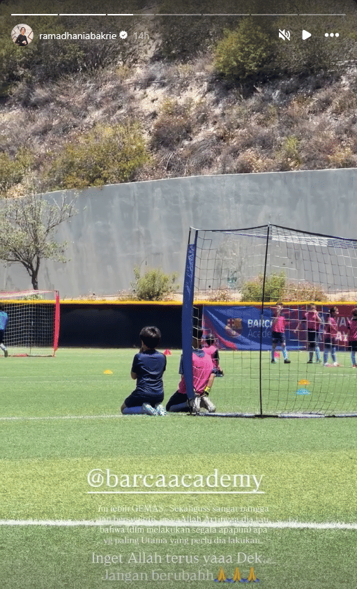 Seorang anak mengenakan seragam latihan sepak bola berwarna biru berlutut di lapangan rumput Barcelona Academy bersama anak-anak lain.