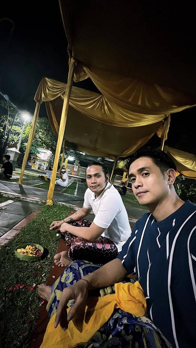 Dua pria duduk bersila di bawah tenda berwarna kuning saat perayaan Kuningan, mengenakan pakaian tradisional Bali pada malam hari.