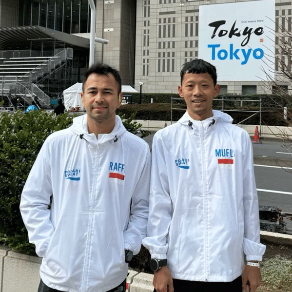 Dua pria mengenakan jaket putih bertuliskan nama mereka berdiri di luar gedung dengan papan bertuliskan Tokyo di latar belakang.