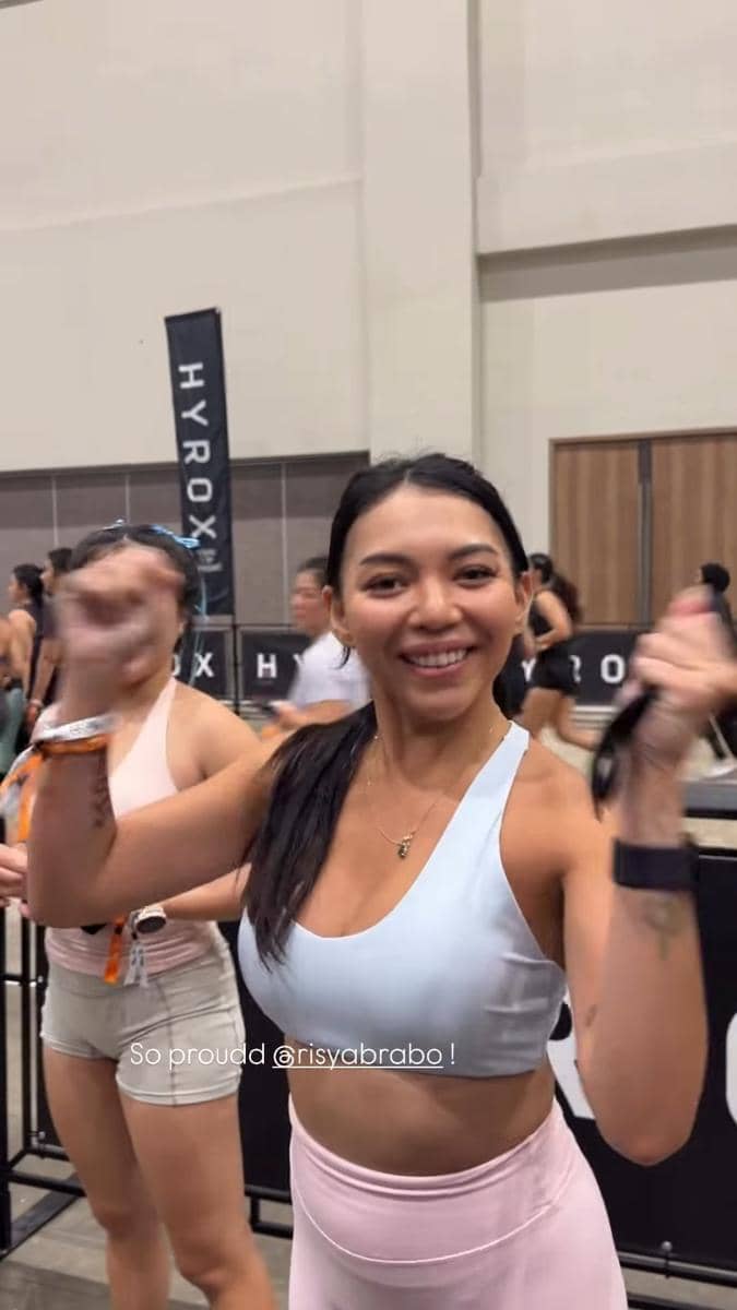 Seorang peserta wanita tersenyum dan mengangkat tangan dengan semangat di acara olahraga HYROX Jakarta 2026 di dalam ruangan.