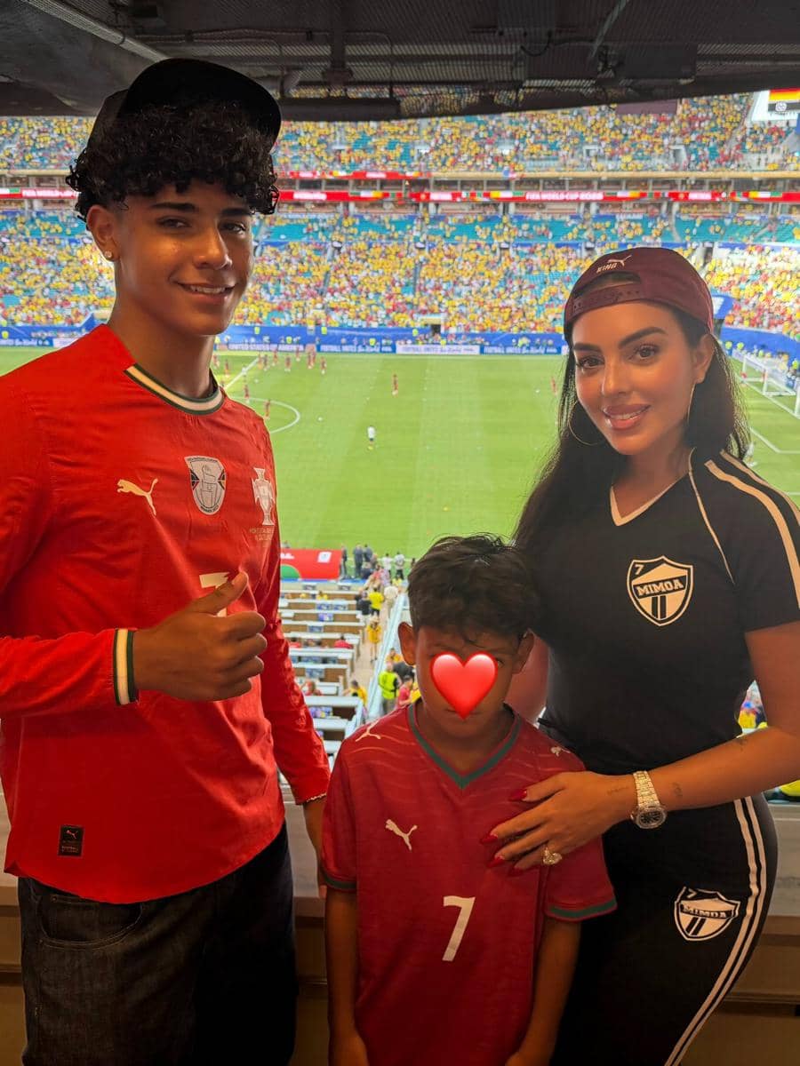 Seorang wanita dan dua anak laki-laki mengenakan jersey sepak bola berpose di tribun stadion saat pertandingan Piala Dunia berlangsung.