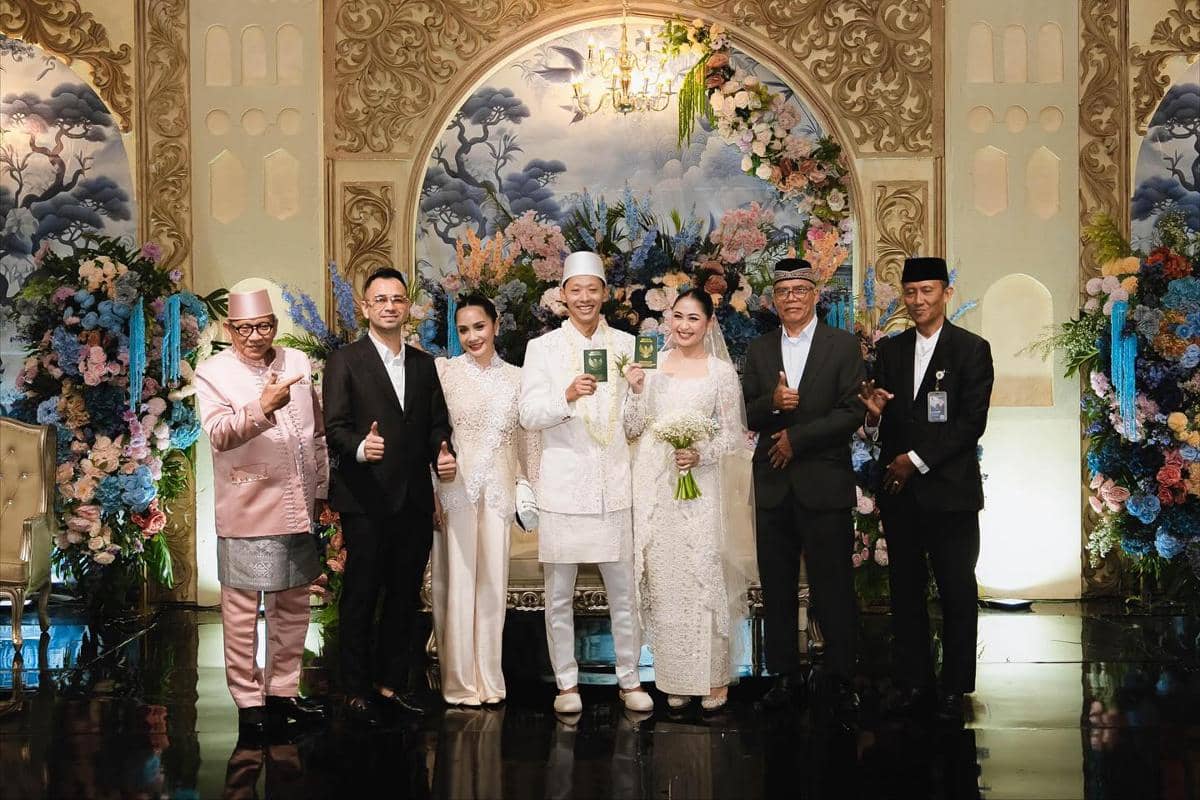Pasangan pengantin berbusana adat putih berpose bersama keluarga dan tamu di pelaminan berhias bunga warna-warni pada acara pernikahan.