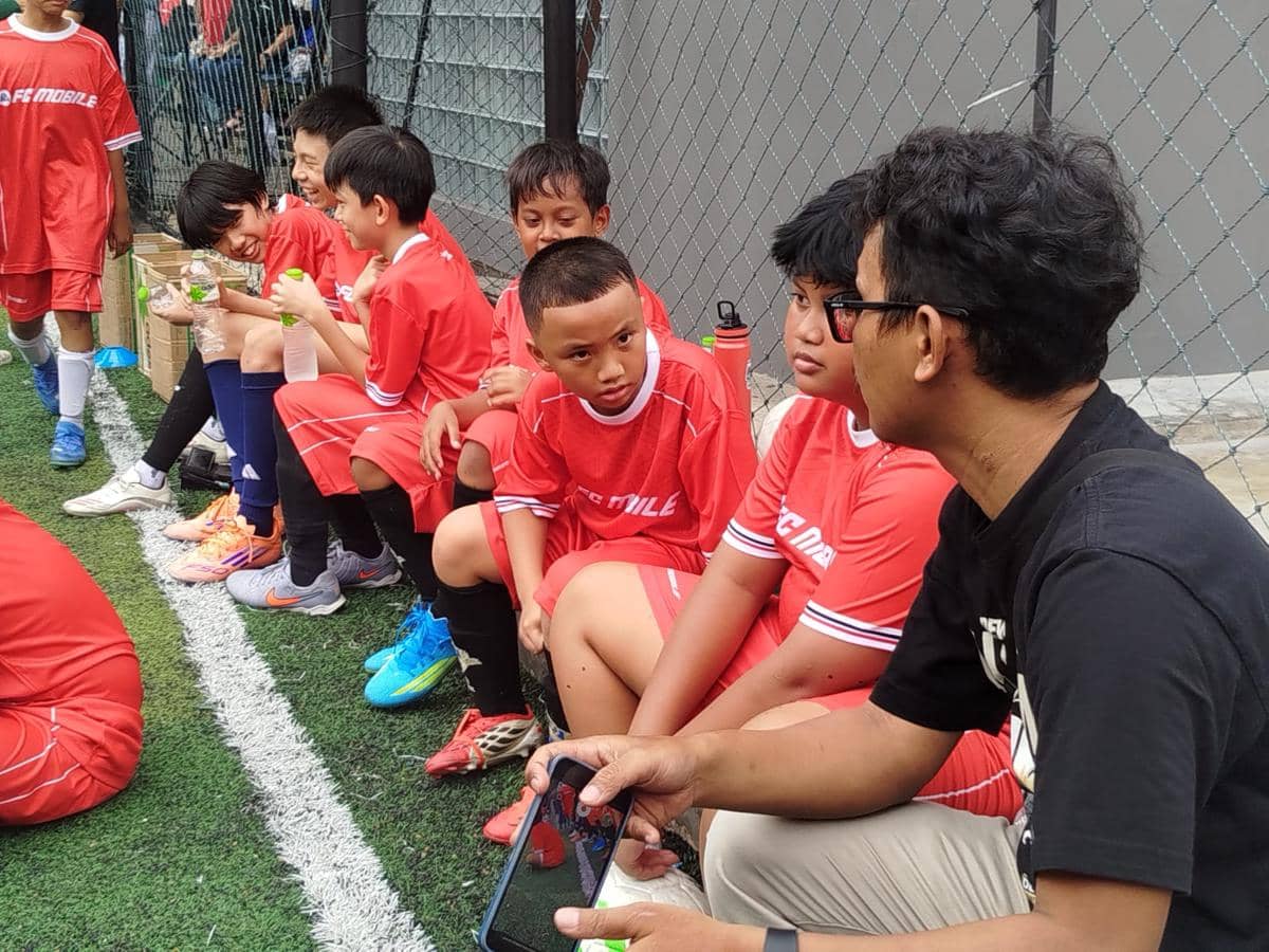 Seorang pelatih berbicara dengan anak-anak yang mengenakan seragam sepak bola merah di pinggir lapangan sintetis.
