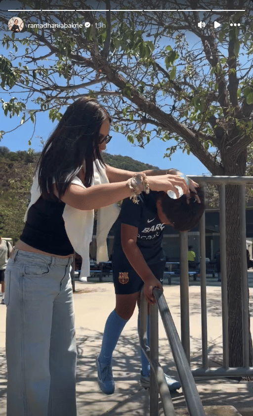 Seorang wanita membantu anak laki-laki yang mengenakan seragam sepak bola Barcelona Academy di area luar dengan pepohonan rindang.