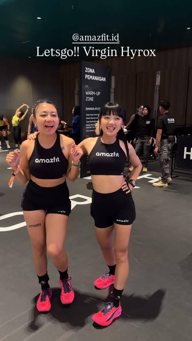 Dua peserta wanita mengenakan pakaian olahraga hitam bertuliskan Amazfit tersenyum dan berpose di area pemanasan acara Hyrox Jakarta 2026.