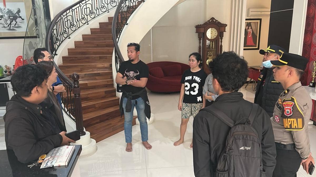 Beberapa petugas kepolisian berbicara dengan sejumlah orang di dalam rumah berlantai kayu dengan tangga melingkar dan jam besar di sudut ruangan.