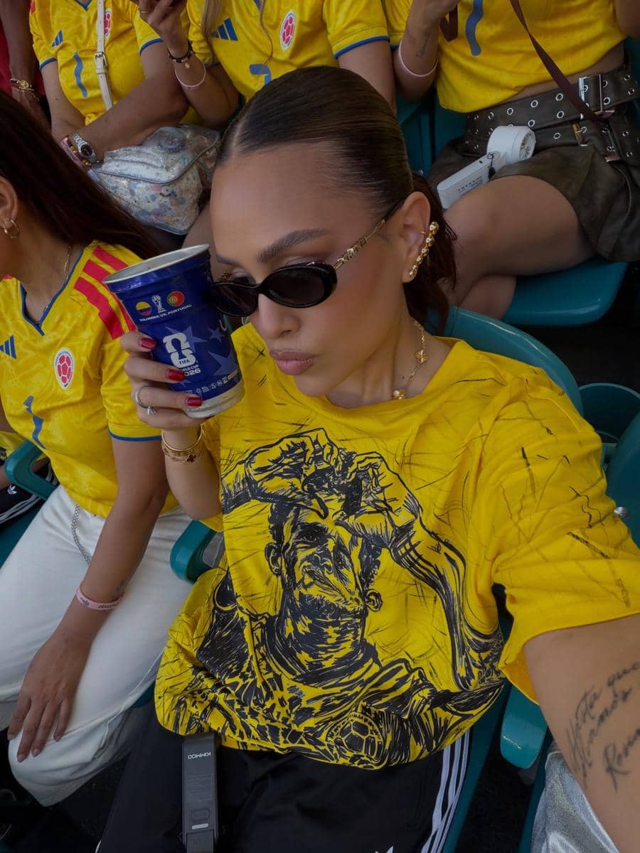 Seorang wanita mengenakan jersey kuning tim nasional Kolombia berpose sambil memegang gelas minuman di tribun stadion bersama pendukung lain.