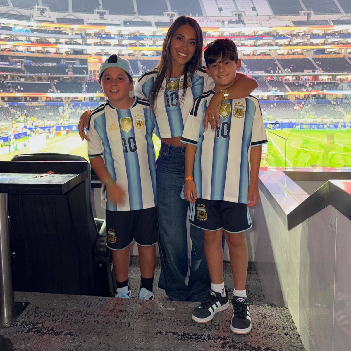 Seorang wanita bersama dua anak mengenakan jersey tim nasional Argentina berdiri di tribun stadion saat pertandingan berlangsung.