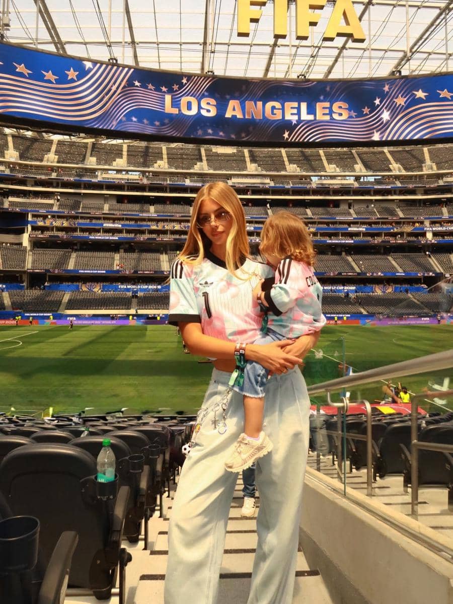 Seorang wanita mengenakan jersey sepak bola memegang anak kecil di tribun stadion Los Angeles dengan latar tulisan FIFA di atasnya.