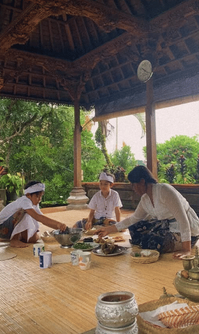 Keluarga mengenakan pakaian adat Bali duduk bersama di bale bambu terbuka sambil menyiapkan sesajen untuk perayaan Kuningan.