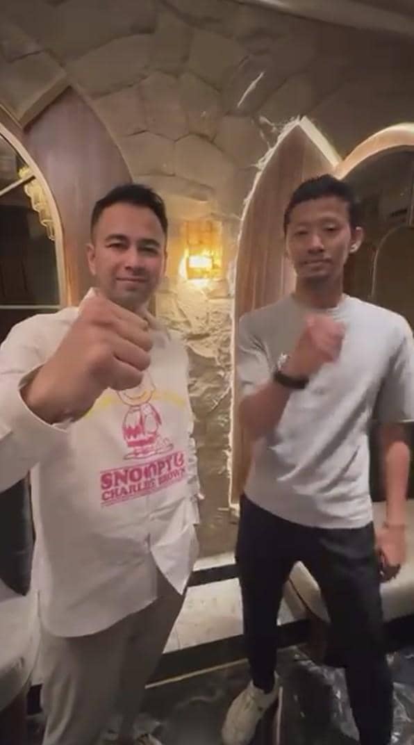 Dua pria berdiri berdampingan di dalam ruangan dengan dinding batu dan pencahayaan hangat, keduanya mengangkat tangan dengan gestur salam.