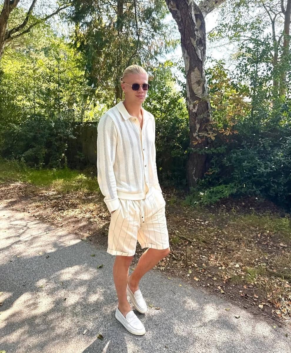 Seorang pria berambut pirang mengenakan kemeja rajut putih, celana pendek bergaris, dan sepatu slip-on putih berjalan di jalan setapak teduh.