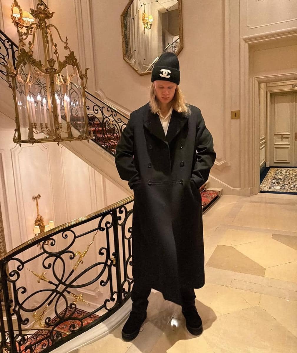 Seorang pria berambut panjang mengenakan mantel hitam panjang dan topi beanie hitam berdiri di tangga dengan interior mewah bergaya klasik.