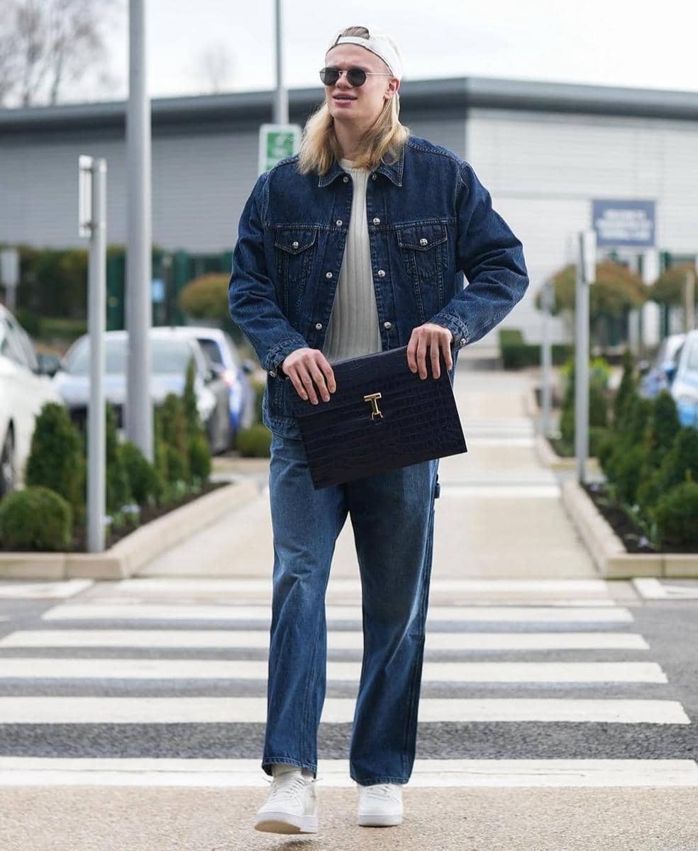 Seorang pria berambut panjang mengenakan jaket denim biru, celana jeans, dan topi putih sambil membawa tas tangan hitam di area parkir.