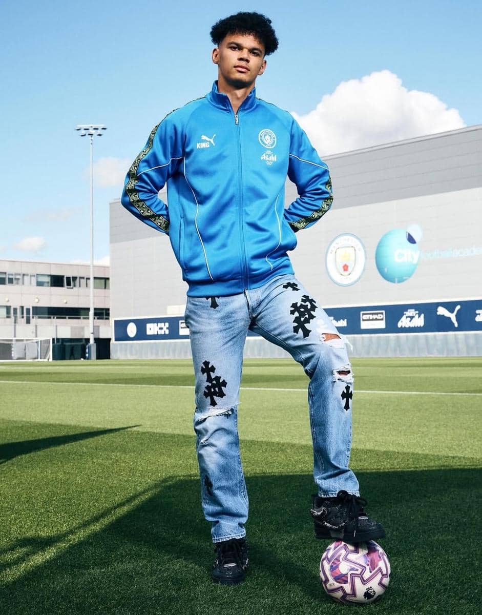 Seorang pria mengenakan jaket biru dan celana jeans robek berdiri di lapangan sepak bola dengan satu kaki di atas bola.