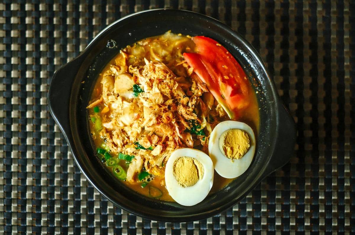 Semangkuk bubur dengan topping ayam suwir, telur rebus, irisan tomat, dan taburan bawang goreng di atas meja anyaman.