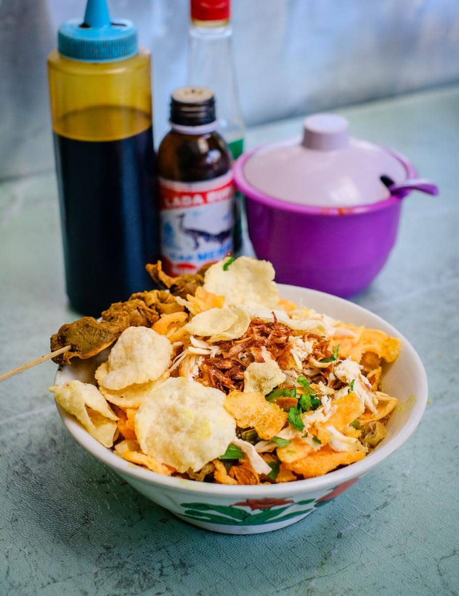 Semangkuk bubur ayam dengan topping kerupuk, suwiran ayam, dan bawang goreng disajikan di meja bersama botol saus dan lada cair.