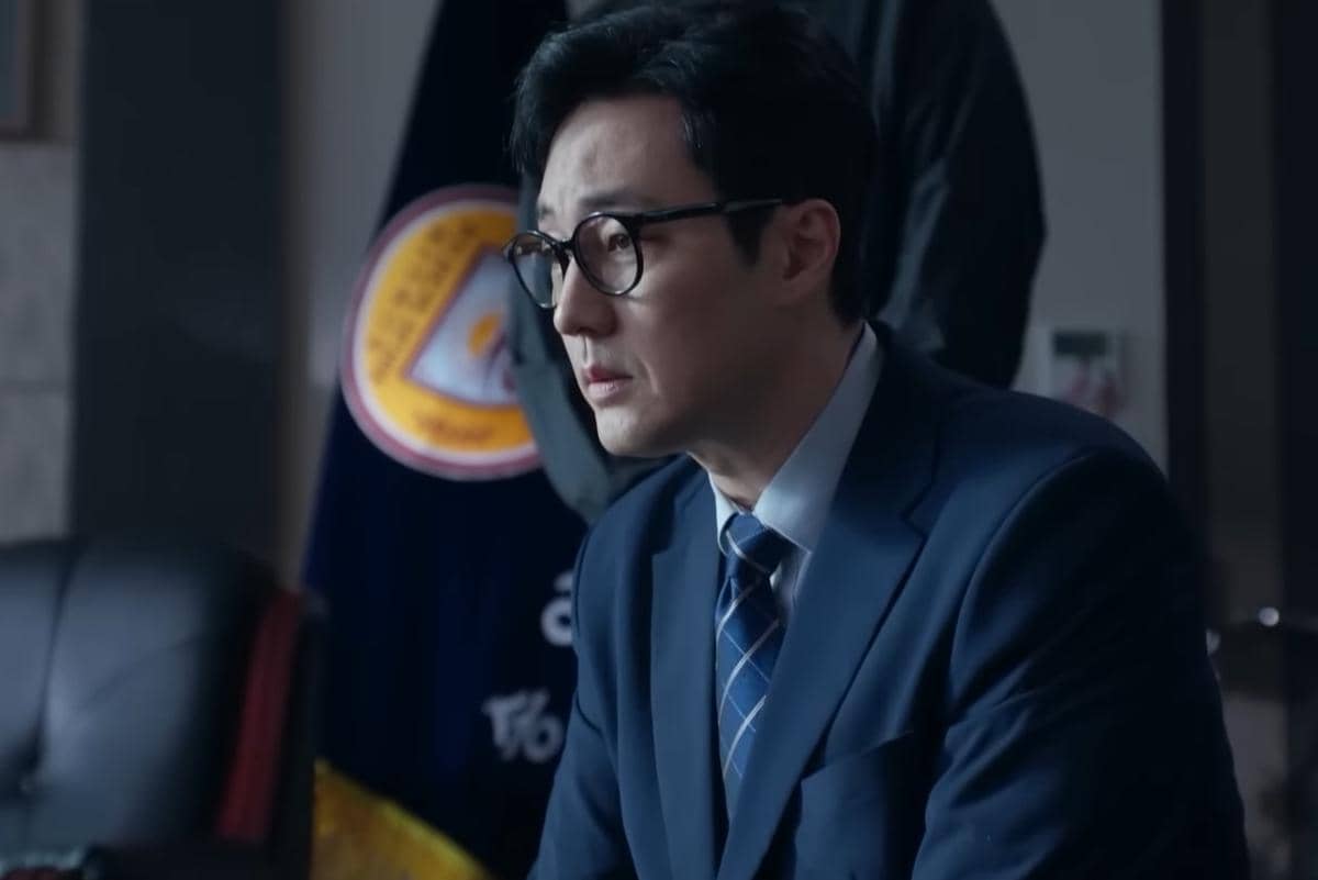 Seorang pria mengenakan setelan jas biru dan kacamata duduk di ruang kantor dengan ekspresi serius dalam cuplikan drama Agent Kim Reactivated.