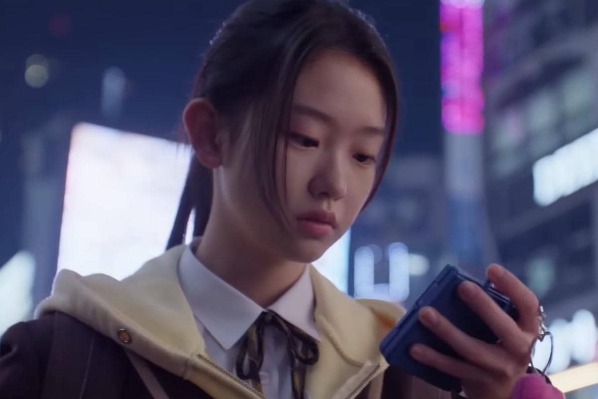 Seorang gadis muda mengenakan seragam sekolah dan jaket krem sedang menatap ponsel di tengah suasana malam kota yang bercahaya neon.
