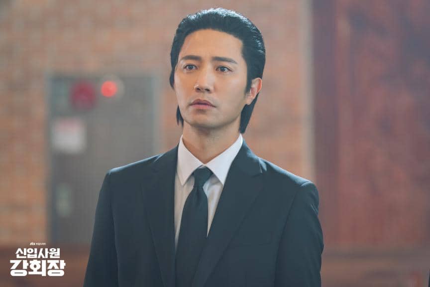 Seorang pria mengenakan setelan jas hitam dan dasi berdiri di dalam ruangan dengan latar dinding bata dalam adegan drama Korea Reborn Rookie.