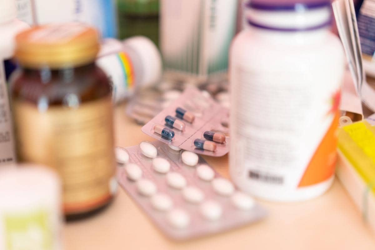 Beberapa botol suplemen dan kemasan obat kapsul serta tablet tersusun di atas meja dengan latar belakang buram.