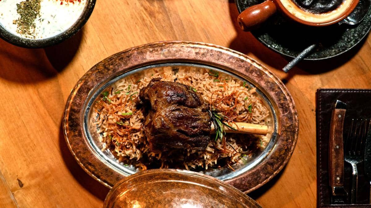 Nasi kebuli dengan potongan daging kambing besar disajikan di atas piring logam oval di meja kayu bersama hidangan pendamping.