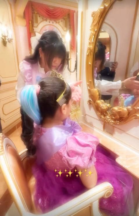 Seorang anak kecil mengenakan gaun dan mahkota duduk di depan cermin berhias emas di Disneyland, dibantu oleh petugas berdandan.