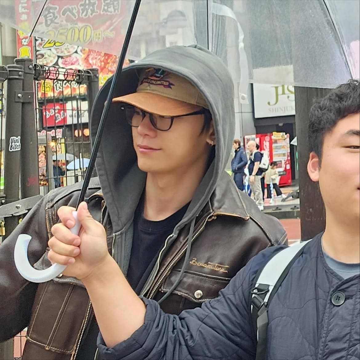 Seorang pria mengenakan jaket kulit, hoodie abu-abu, topi cokelat, dan kacamata sambil memegang payung transparan di jalan kota.