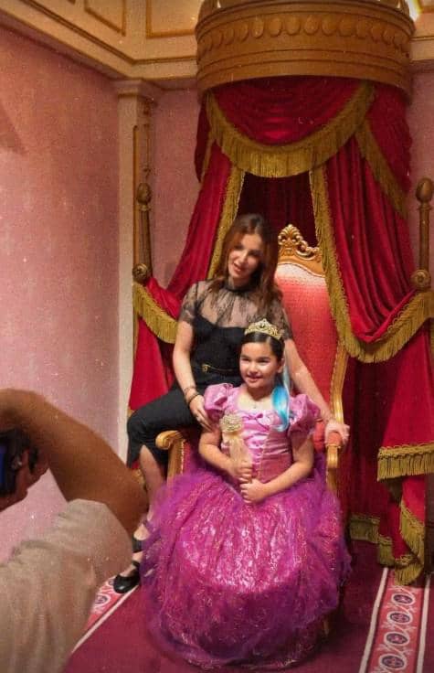 Seorang anak mengenakan gaun putri berwarna ungu duduk di kursi takhta bersama seorang wanita di ruangan bertema kerajaan di Disneyland.