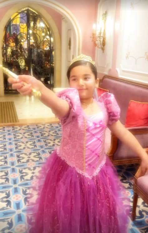 Seorang anak kecil mengenakan gaun putri berwarna ungu muda dengan mahkota emas dan tongkat di tangan, berpose di dalam ruangan bergaya istana.