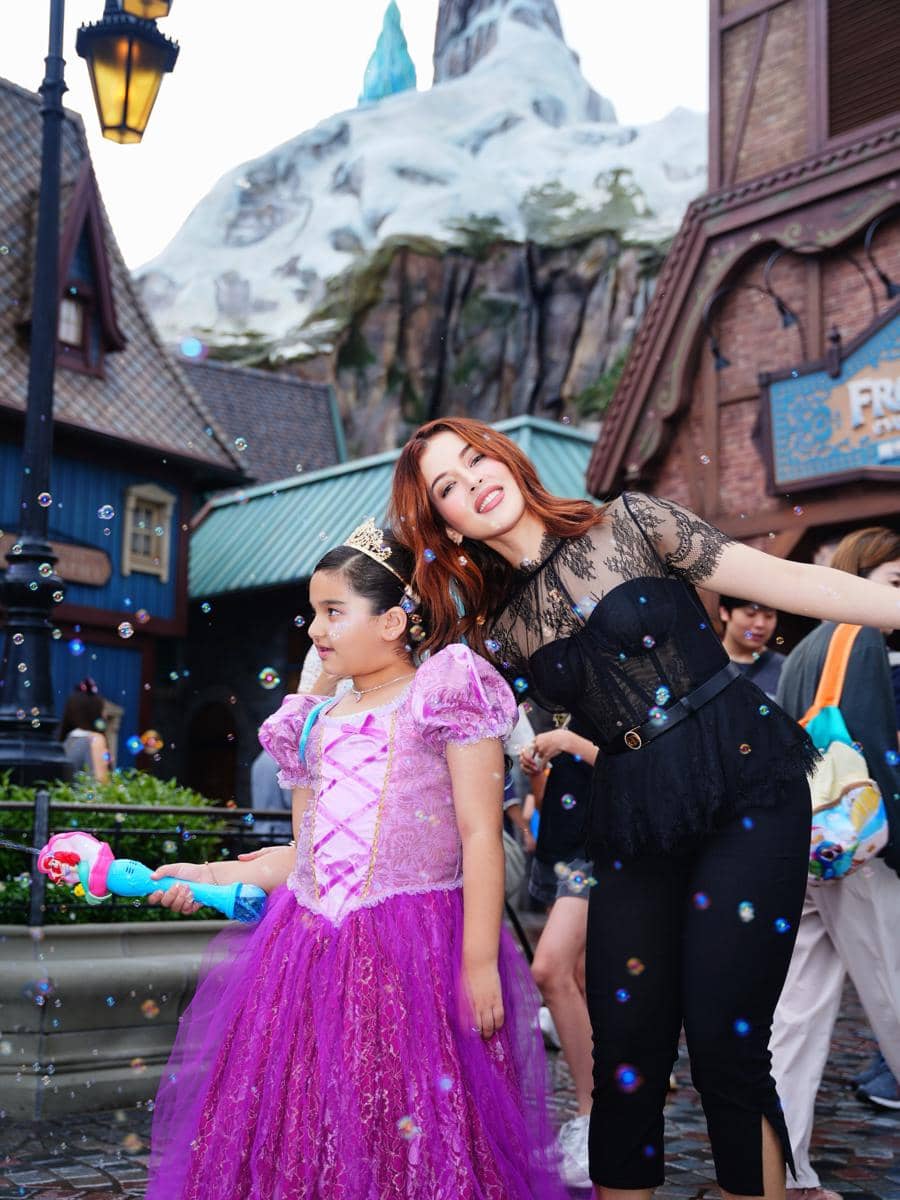 Seorang anak mengenakan gaun ungu bergaya putri bersama seorang wanita di area bertema dongeng Disneyland dengan latar gunung es buatan.