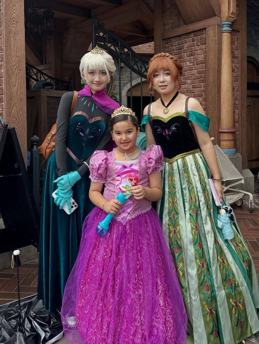 Seorang anak mengenakan gaun ungu ala putri bersama dua perempuan berkostum karakter Frozen di area bertema kastel Disneyland.