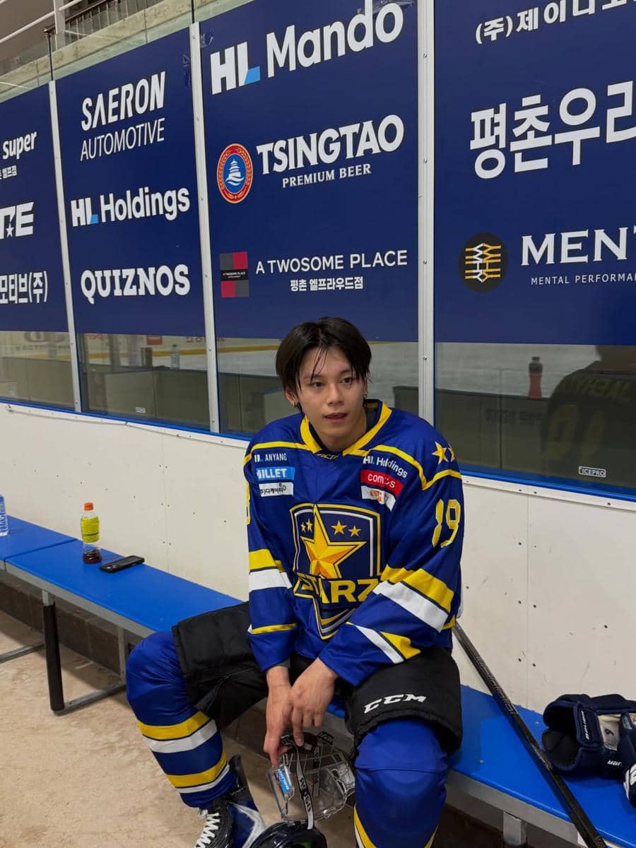 Seorang pemain hoki es duduk di bangku pemain dengan seragam biru dan kuning, memegang tongkat hoki di arena latihan.