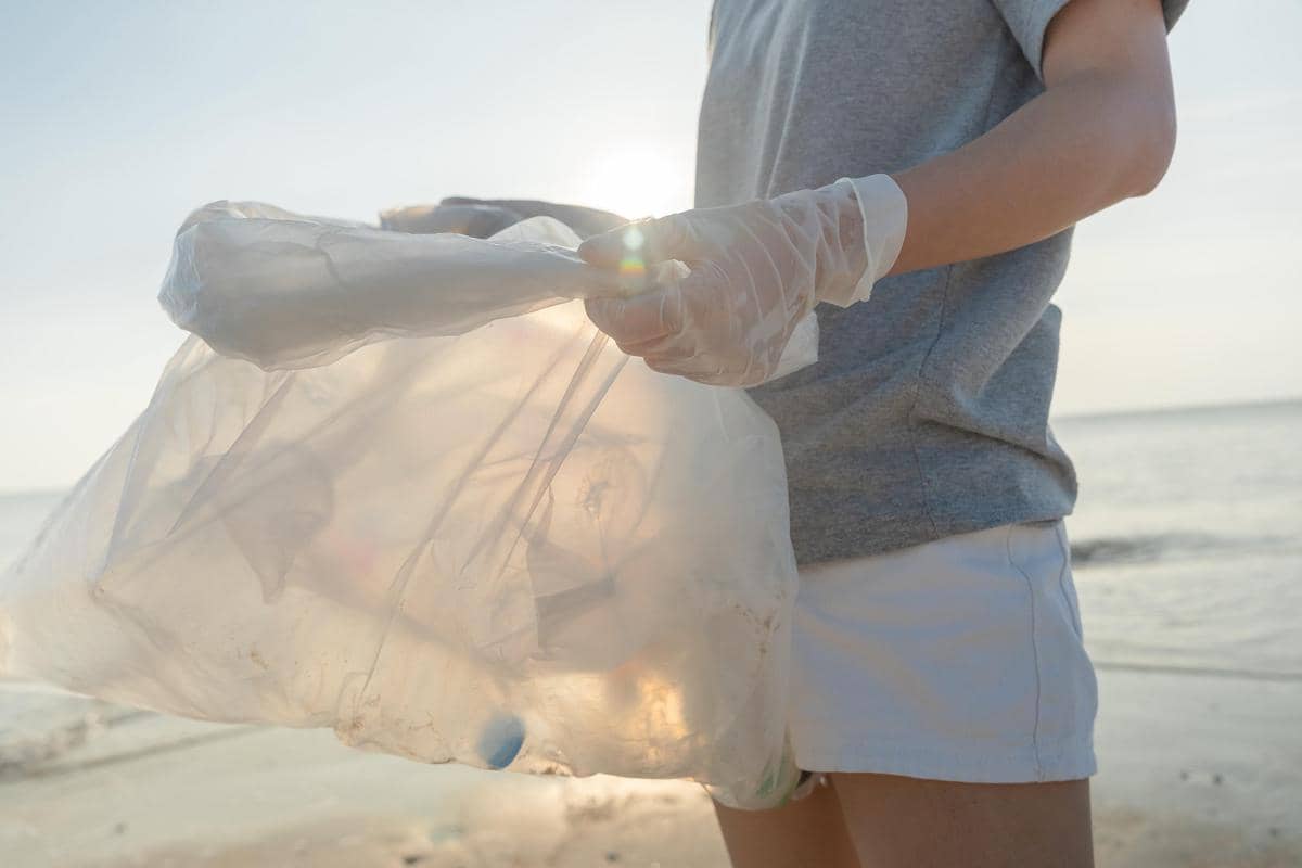 Seseorang mengenakan sarung tangan plastik memegang kantong plastik berisi sampah di tepi pantai saat matahari terbenam.