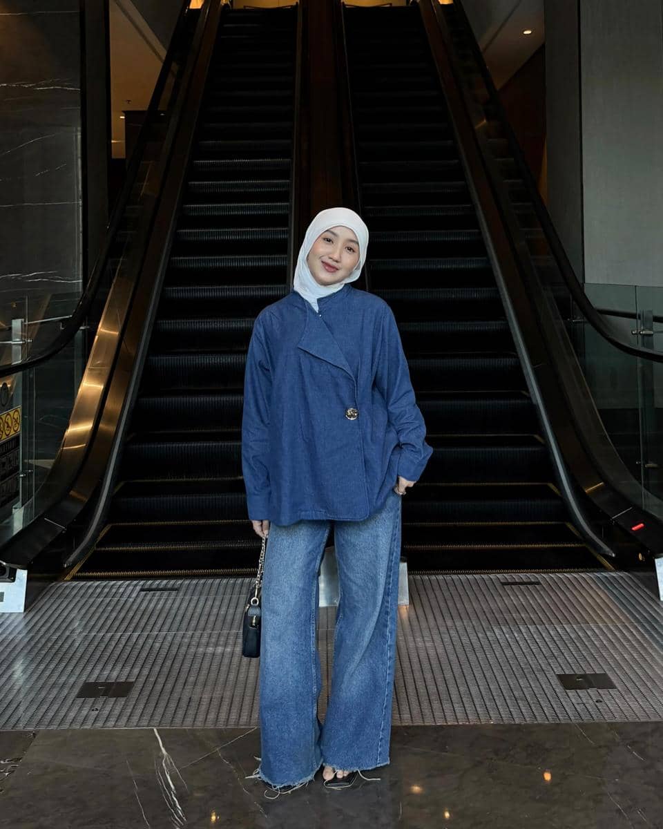 Perempuan berhijab putih mengenakan atasan denim longgar dan celana jeans lebar berpose di depan eskalator di area gedung modern.