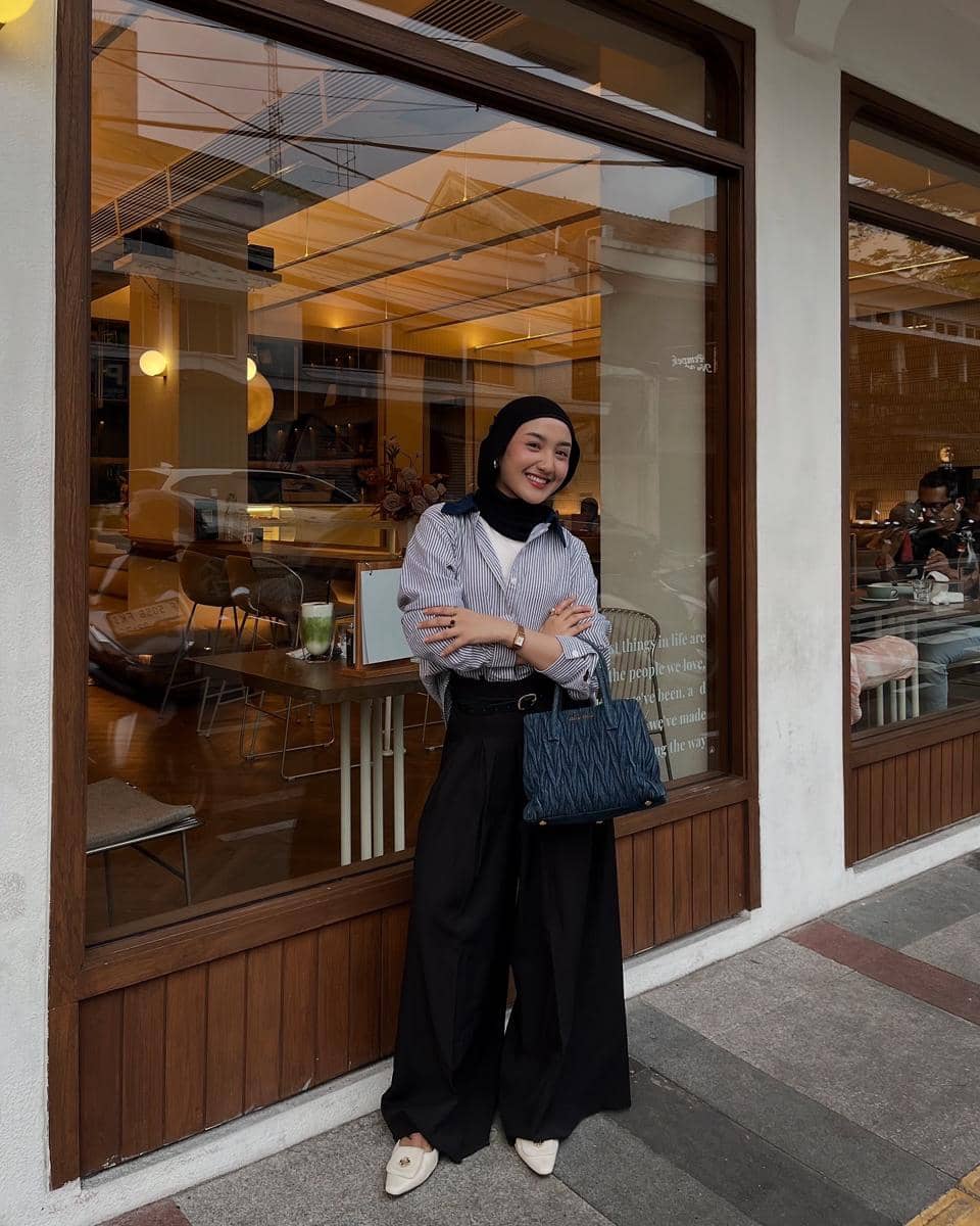 Seorang wanita berhijab mengenakan kemeja bergaris dan celana hitam lebar sambil berpose di depan kafe dengan jendela besar.