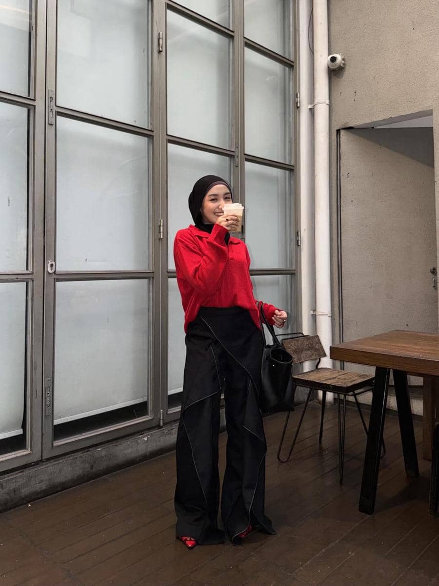 Seorang wanita berhijab mengenakan kemeja merah dan celana hitam longgar sambil memegang minuman di area kafe dengan jendela besar.