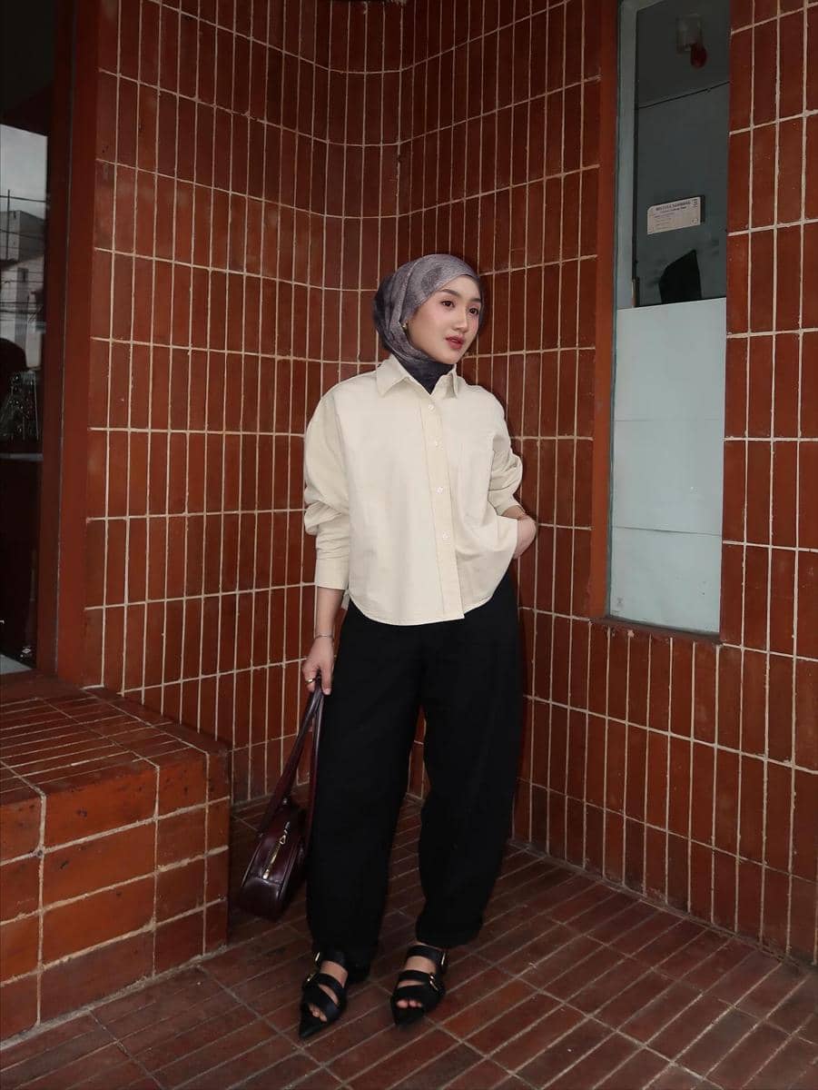 Seorang wanita berhijab mengenakan kemeja krem dan celana hitam longgar sambil membawa tas cokelat di depan dinding bata cokelat.