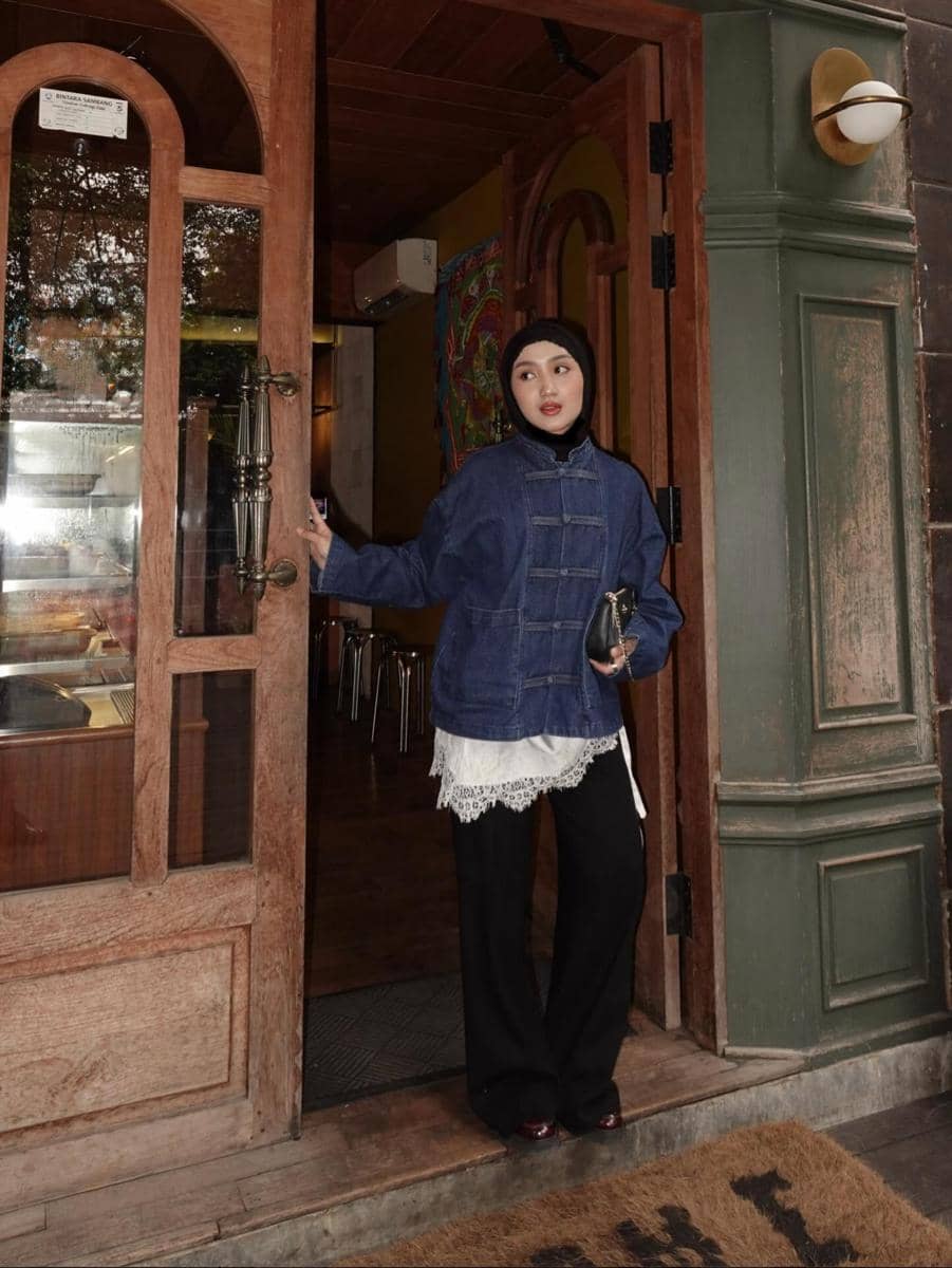 Seorang wanita berhijab mengenakan jaket denim biru, blouse renda putih, dan celana hitam berdiri di pintu masuk kafe bergaya kayu.