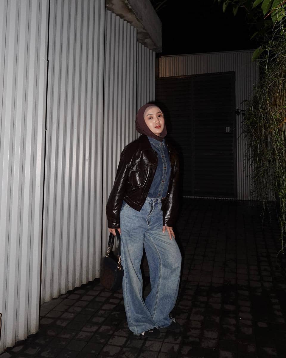 Seorang wanita berhijab mengenakan jaket kulit cokelat, kemeja denim, dan celana jeans longgar sambil berpose di area outdoor malam hari.