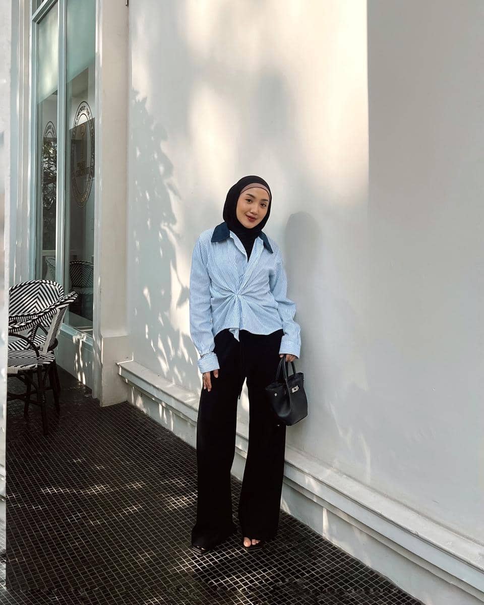 Seorang wanita berhijab mengenakan kemeja biru bergaris dan celana panjang hitam sambil memegang tas hitam di area luar ruangan.